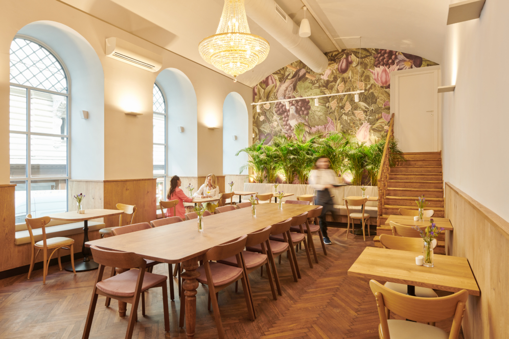 Helles, modernes Café mit Holztischen und -stühlen, großen Rundbogenfenstern, einer mit Blumen verzierten Wand, Topfpflanzen und einem Kronleuchter. Drei Personen sitzen an einem Tisch, während eine Bedienung vorbeigeht.