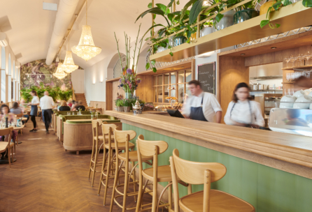Ein stilvolles, modernes Restaurant mit Holzstühlen an einer langen Bar, Kronleuchtern, grünen Akzenten, üppigen Pflanzen und bewegten Mitarbeitern hinter dem Tresen. Die Gäste sitzen an Tischen im Hintergrund.