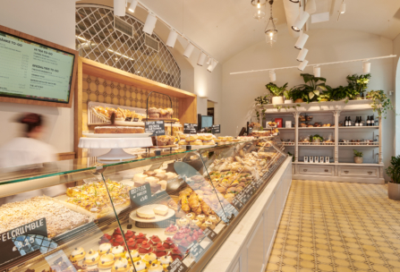 Eine helle Bäckerei mit einer Glasauslage, die mit verschiedenen Backwaren, Kuchen und Desserts gefüllt ist. In den Regalen im Hintergrund stehen Pflanzen und Produkte. Warme Beleuchtung und geflieste Böden schaffen eine einladende Atmosphäre.