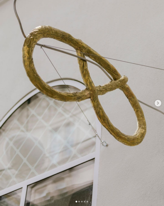 Eine große goldene, brezelförmige Skulptur hängt an Drähten über einem Gebäudeeingang mit einer Glastür und einem Rundbogenfenster.