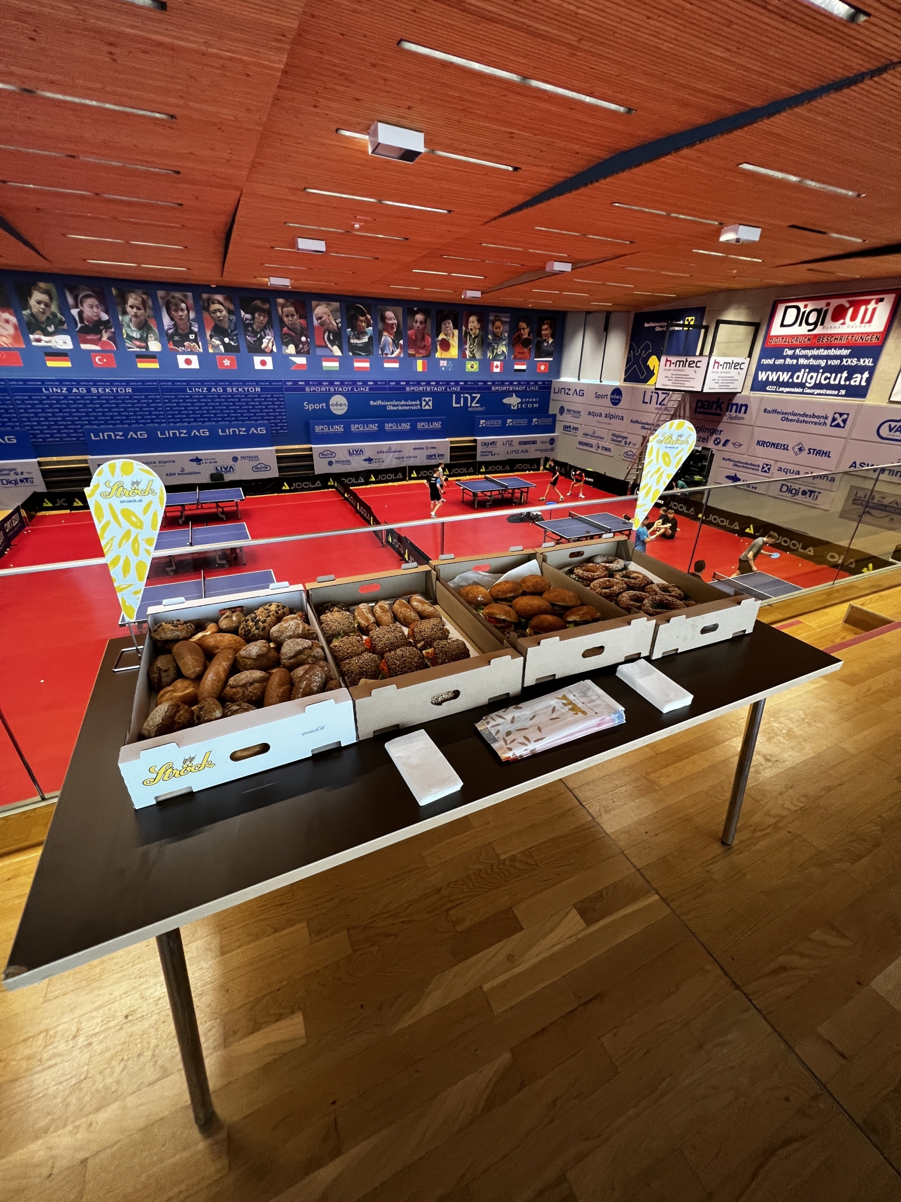 Im Vordergrund einer Sporthalle mit rotem Bodenbelag und Bannern an den Wänden steht ein Tisch mit verschiedenen Backwaren in Tabletts. Im Hintergrund sind die Sitzplätze und der Veranstaltungsbereich zu sehen.