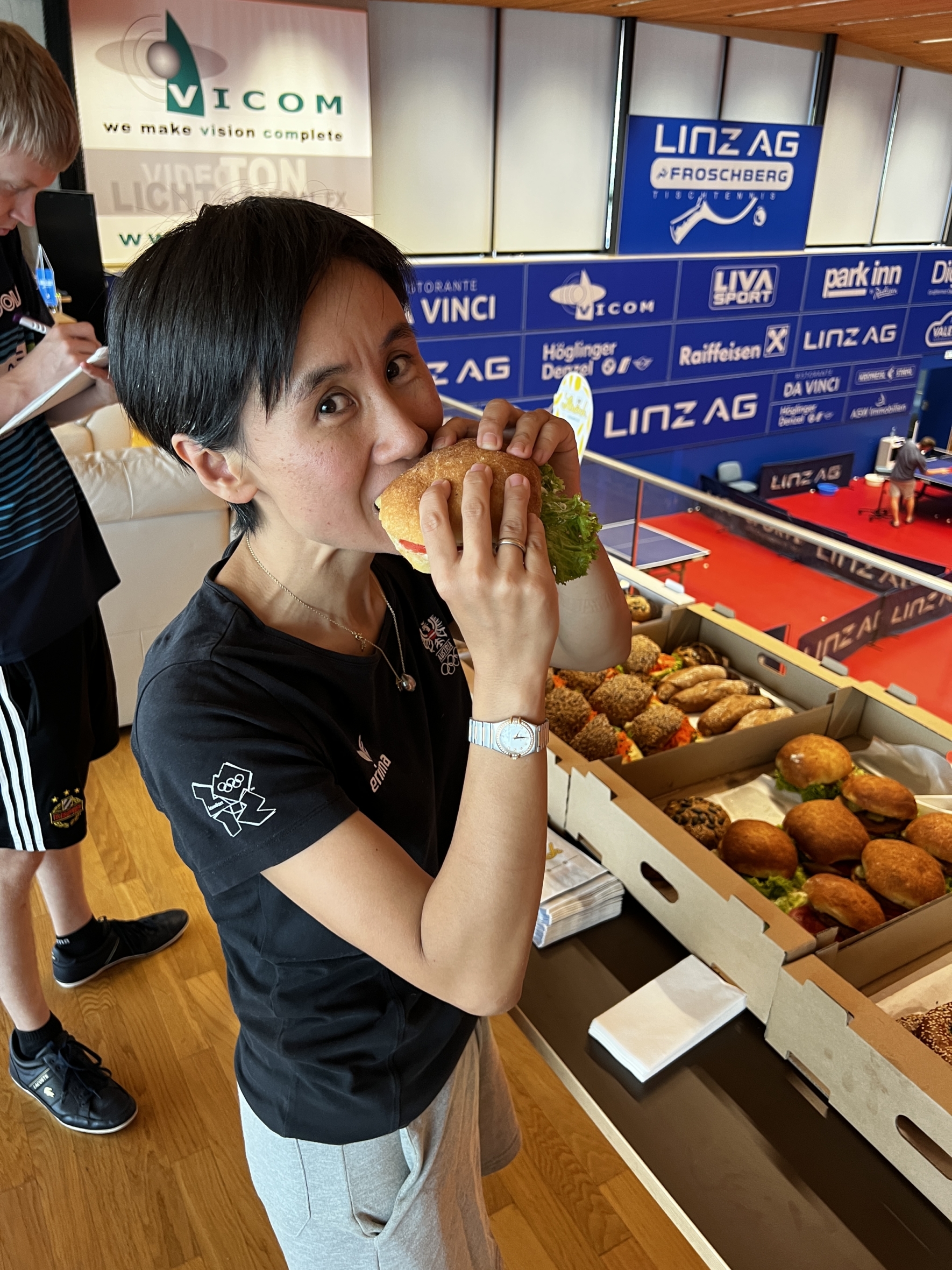 Eine Frau lächelt, während sie ein großes Sandwich in der Hand hält und isst, während sie in der Nähe von Tischen steht, die mit weiteren Sandwiches gefüllt sind. Sie ist in einem Raum mit Sportbannern und einer weiteren Person im Hintergrund zu sehen.