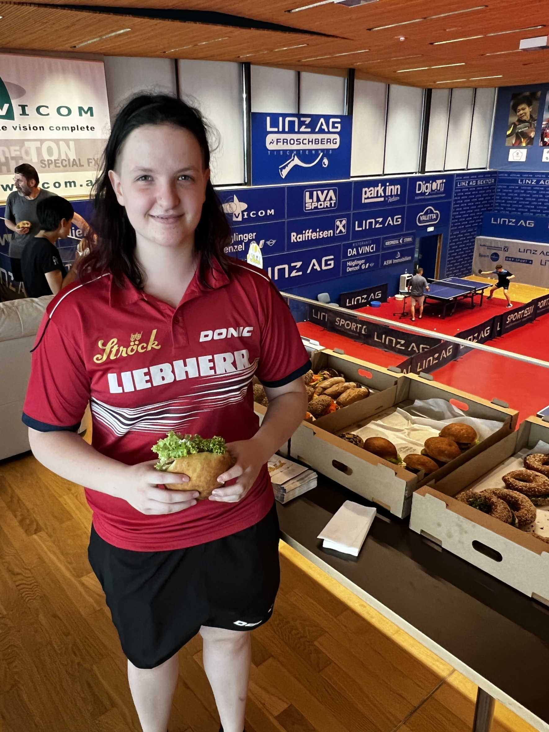 Eine Frau in einem roten Sporttrikot steht in einem Raum und hält ein Sandwich mit Salat in der Hand. Hinter ihr stehen Tische mit Brötchen und ein Hintergrund mit Logos und einer Tischtennisplatte.
