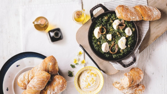 Ein Tisch mit einer Pfanne mit gebackenen Schnecken in grüner Soße, übergossen mit Sahne, umgeben von knusprigen Brötchen, Olivenöl, Balsamico-Essig und einer Schale mit cremigem Dip mit Zitronenschale.