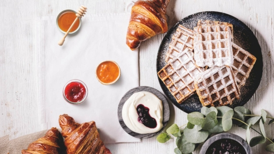Ein Frühstücksbuffet mit Waffeln, die mit Puderzucker bestäubt sind, Croissants, kleinen Schälchen mit Marmelade, Honig und Sahne und Eukalyptuszweigen auf einem hellen Holztisch.