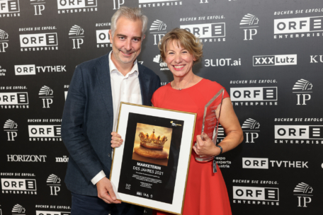 Zwei Personen stehen lächelnd vor einer Markenkulisse. Die Frau in einem roten Kleid hält eine gläserne Trophäe und eine gerahmte Auszeichnung "Marketerin des Jahres 2021" mit einem Kronenbild. Der Mann neben ihr trägt eine Anzugsjacke und ein Hemd.