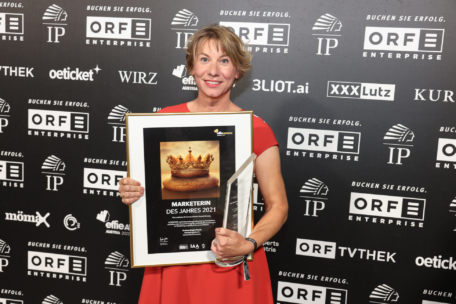 Eine Frau in einem roten Kleid hält eine gerahmte Auszeichnung "Marketerin des Jahres 2021" und eine Glastrophäe in der Hand und steht vor einem schwarzen Hintergrund mit verschiedenen Sponsorenlogos.