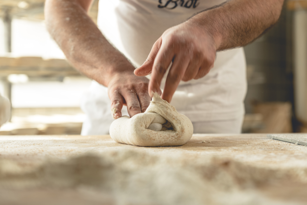 Die Hände einer Person kneten und formen Teig auf einer bemehlten Fläche, was auf die Zubereitung von Brot oder Gebäck in einer Bäckerei oder Küche hindeutet. Im Hintergrund sind Regale mit weiteren Backwaren zu sehen.