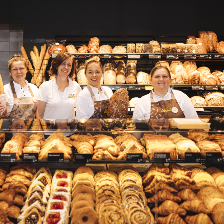 Vier Bäckereiangestellte stehen hinter einer mit Gebäck und Brot gefüllten Glastheke und lächeln in die Kamera. Hinter ihnen stehen Regale mit einer Vielzahl von Broten und Baguettes. Die Bäckerei ist hell erleuchtet und gut organisiert.