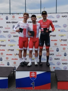 Drei männliche Radrennfahrer in Rennkleidung stehen auf einem Podium, lächeln und halten Auszeichnungen in der Hand. Zwei tragen polnische Trikots (2., 1. Platz), einer trägt ein österreichisches Trikot (3. Platz). Auf einem Hintergrund sind Sponsorenlogos und Veranstaltungshinweise zu sehen.