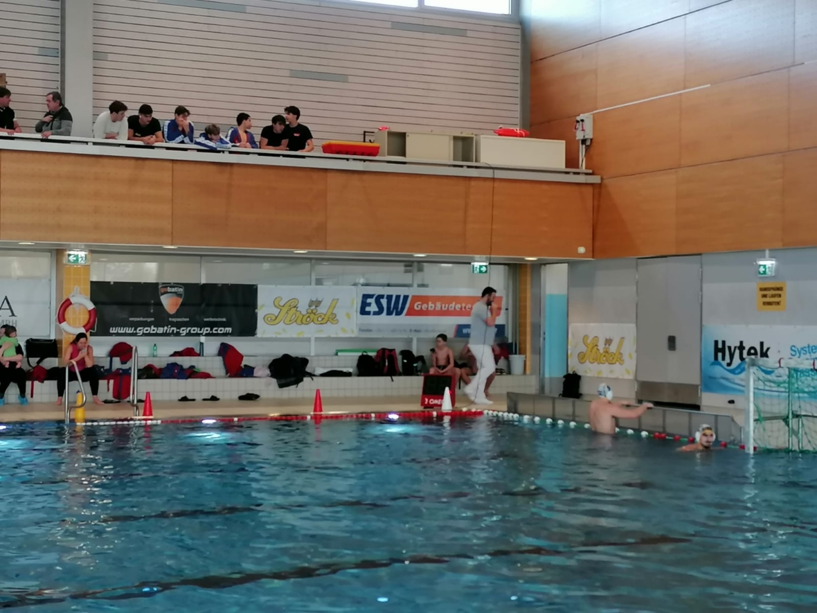 Ein Wasserballspiel in einem Hallenbad; ein Spieler bereitet sich darauf vor, auf das Tor zu schießen, während der Torwart bereit ist. Die Zuschauer sitzen auf den Tribünen darüber, und an der Wand sind Werbebanner zu sehen.