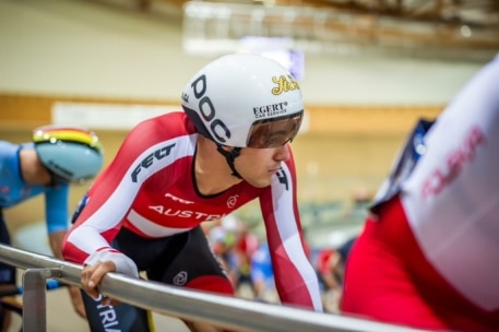 Ein Radfahrer mit weißem Helm und rot-weißer österreichischer Uniform hält sich an einem Geländer fest und bereitet sich auf ein Rennen auf einer Indoor-Radrennbahn vor, mit anderen Radfahrern im Hintergrund.