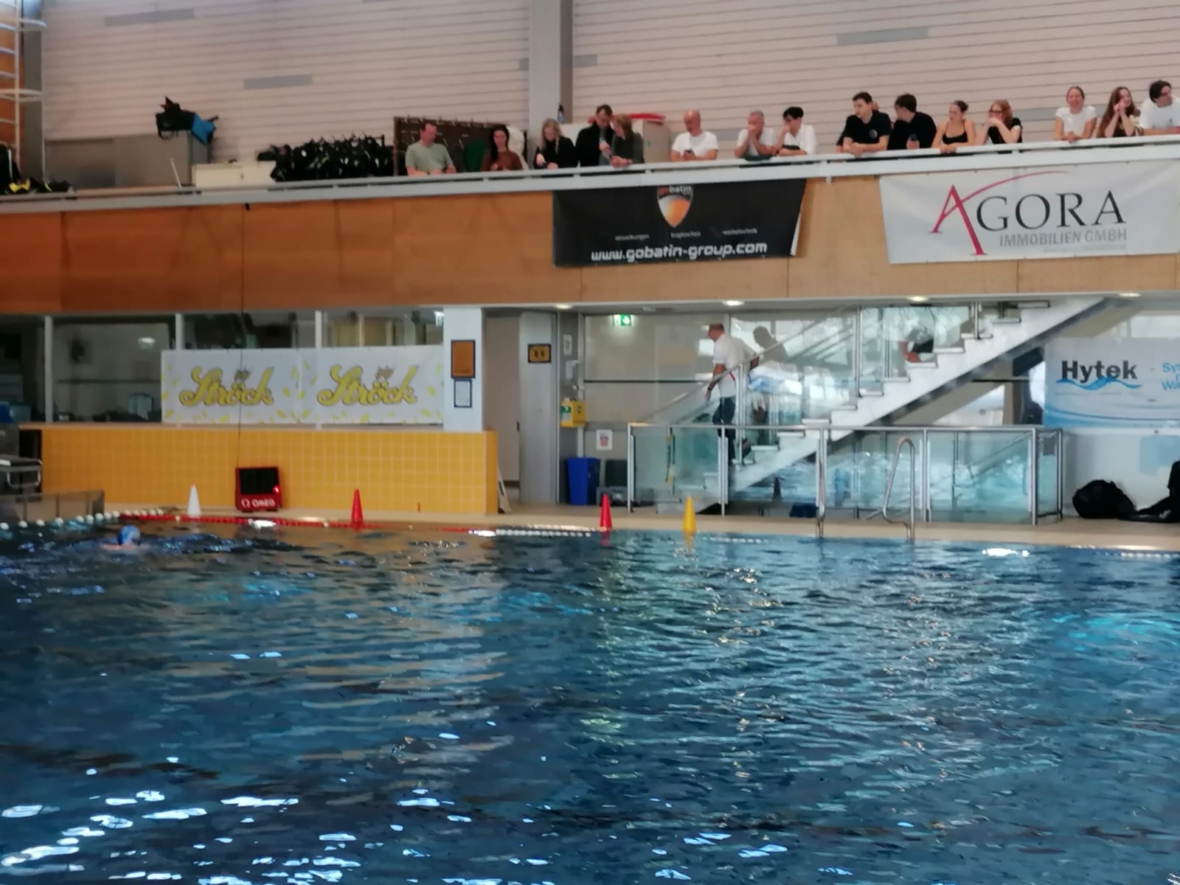 Im Vordergrund ist ein Schwimmbad mit klarem, blauem Wasser zu sehen. Zuschauer sitzen auf Bänken darüber und schauen zu. Es gibt Banner und Schilder an der Wand, und eine Person geht in der Nähe der Treppe am Beckenrand.