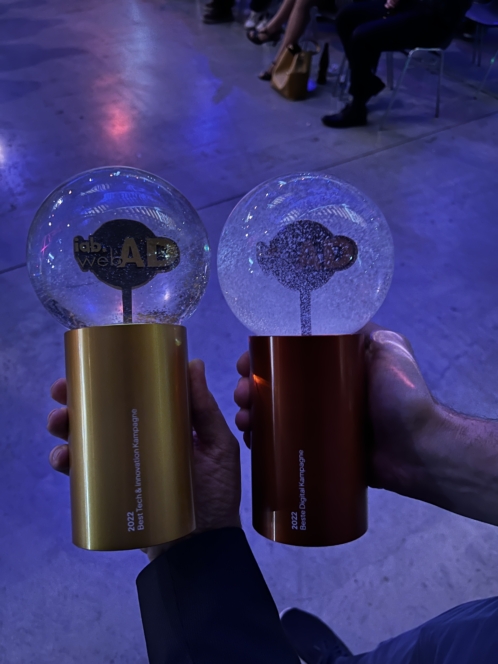 Zwei Hände halten runde, transparente Trophäen mit goldenen und bronzenen Sockeln; die Trophäen sind beschriftet mit "Best in Web3 Native Language" und "Best AI Project Language". Im Hintergrund sitzen Menschen bei einer Veranstaltung.