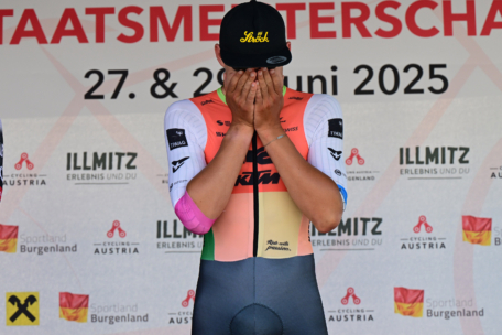 Ein Radfahrer in einem farbenfrohen Rennanzug bedeckt sein Gesicht mit beiden Händen auf einem Podium, vor dem Hintergrund der Veranstaltungsdaten der Österreichischen Radmeisterschaften vom 27. bis 29. Juni 2025.