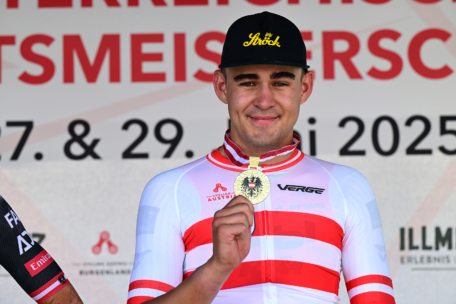 Ein Radfahrer in einem weiß-rot gestreiften Trikot und einer schwarzen Mütze lächelt, während er eine Goldmedaille vor einem Hintergrund mit Logos und Text hochhält.