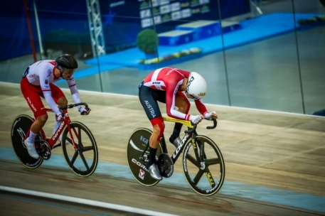 Zwei Radfahrer fahren ein Rennen auf einer Indoor-Velodrom-Bahn. Beide tragen aerodynamische Helme und Rennanzüge; der eine trägt einen rot-weißen Anzug mit "Austria" auf dem Bein, der andere einen rot-weißen Anzug mit "Polska".