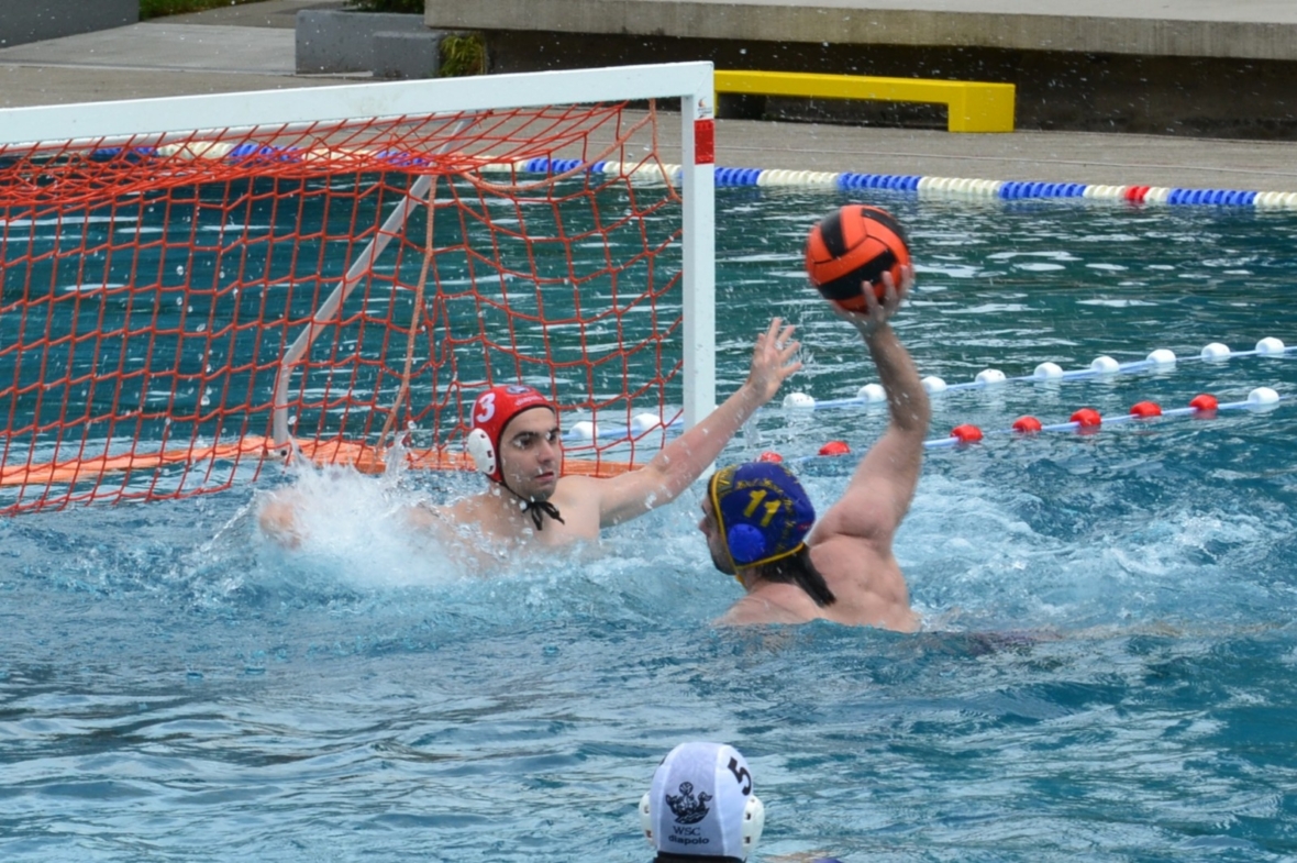 Ein Wasserballspieler mit blauer Mütze bereitet sich darauf vor, den Ball in Richtung Tor zu schießen, während ein Torwart mit roter Mütze versucht, den Ball zu blocken. Beide befinden sich in einem Becken, in dem das Tornetz und die Seile sichtbar sind.