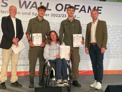 Fünf Personen posieren in einem Innenraum vor einer "Wall of Fame"-Kulisse. Drei in grünen Uniformen halten Urkunden, eine Frau im Rollstuhl hält ebenfalls eine Urkunde, und zwei Männer in Anzügen stehen lächelnd an beiden Enden der Gruppe.