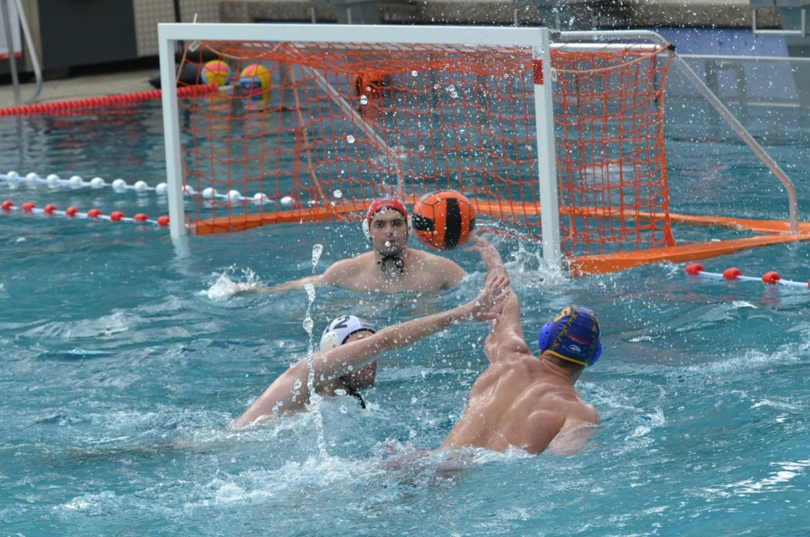 Ein Wasserballspieler mit blauer Mütze wirft den Ball in Richtung Tor, während der Torwart und ein Verteidiger mit weißen Mützen versuchen, den Schuss in einem Schwimmbecken während eines Spiels zu blockieren.
