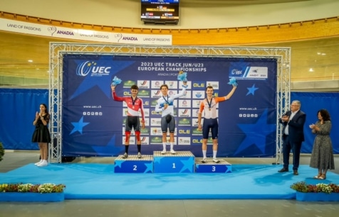 Drei männliche Radsportler stehen bei den UEC-Bahn-Junioren/U23-Europameisterschaften auf einem Podium und nehmen Medaillen entgegen. Sie werden von applaudierenden Offiziellen flankiert, über ihnen ist ein blauer Veranstaltungshintergrund und "Anadia, Portugal" zu sehen.