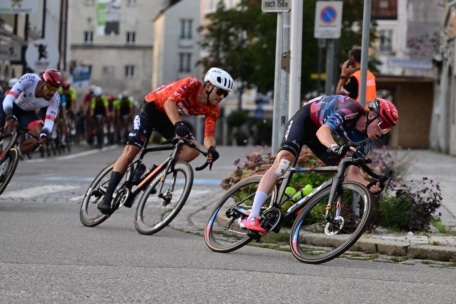 Drei Radfahrer in Helmen und Rennkleidung biegen während eines Radrennens auf einer Stadtstraße scharf in eine Kurve ein, wobei im Hintergrund weitere Radfahrer und Gebäude zu sehen sind.