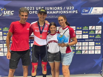 Vier Personen stehen lächelnd vor der Kulisse der UEC Junioren/U23 Bahnradmeisterschaften 2023 zusammen. Der Radsportler in der Mitte trägt ein Österreich-Trikot und eine Medaille und hält Blumen in der Hand, flankiert von drei Unterstützern in Österreich-Trikots.