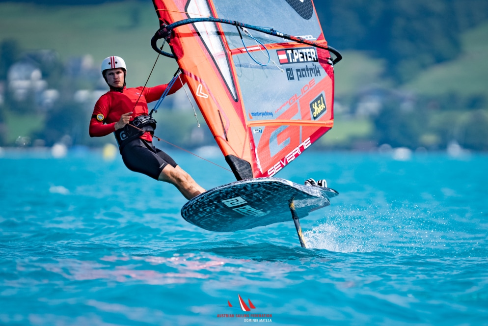 Ein Windsurfer mit rotem Hemd und Helm gleitet auf einem Foilboard über hellblaues Wasser, mit grünen Hügeln und verschwommenen Gebäuden im Hintergrund.