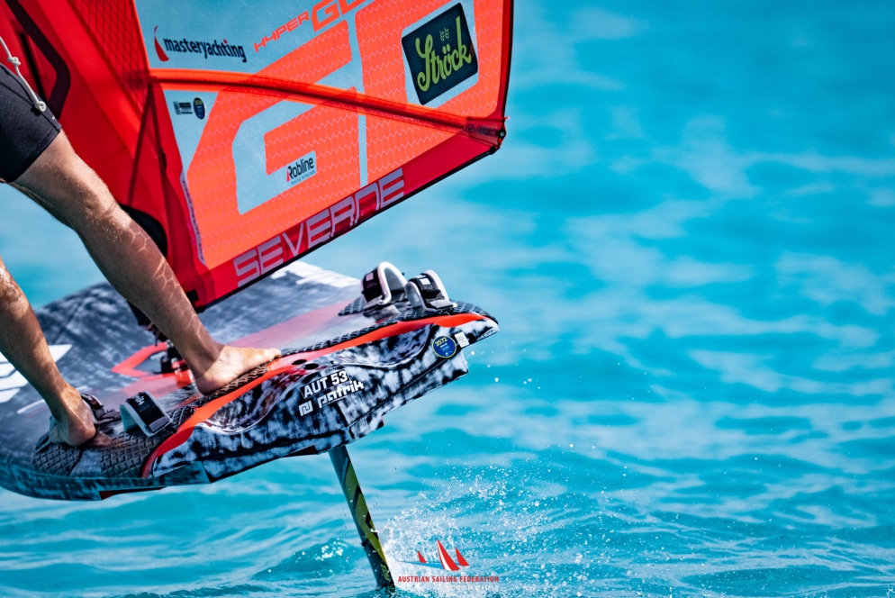Eine Nahaufnahme der Füße einer Person auf einem Foil-Windsurfbrett mit rotem Segel, das über klares blaues Wasser gleitet. Die Finne des Boards schneidet durch das Wasser und verursacht kleine Spritzer.