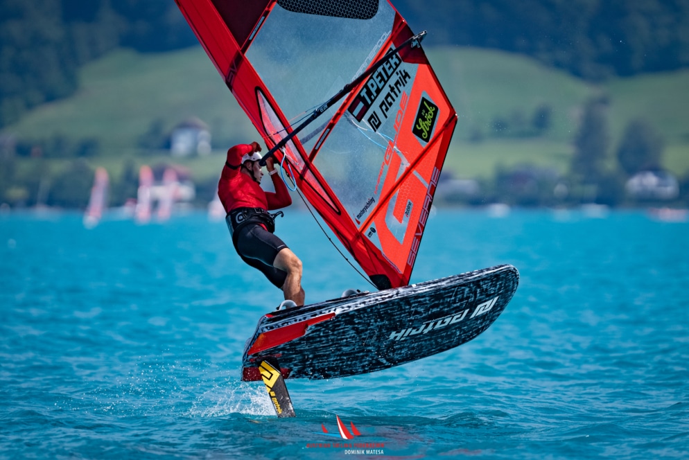 Eine Person in roter Kleidung fährt auf einem Tragflächen-Windsurfbrett über hellblaues Wasser und hebt dabei über die Wasseroberfläche. Grüne Hügel und Häuser sind im verschwommenen Hintergrund unter einem klaren Himmel zu sehen.