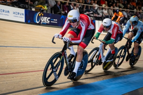 Radfahrer in voller Rennmontur fahren auf einer Indoor-Velodrombahn um die Wette, während im Hintergrund Zuschauer von der Tribüne aus zusehen. Der führende Radfahrer trägt einen rot-weißen Anzug und einen weißen Helm.