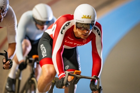 Ein Radfahrer in rot-weißer österreichischer Uniform und weißem Helm führt eine Gruppe auf einer Indoor-Bahn an, zwei Radfahrer folgen dicht dahinter.
