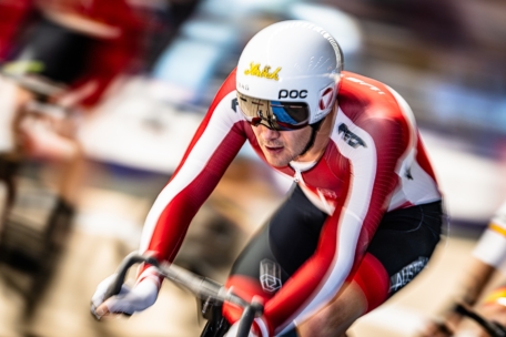 Ein Radfahrer in einem rot-weißen Anzug und mit weißem Helm fährt auf einer Indoor-Bahn, wobei er sich intensiv nach vorne lehnt. Der Hintergrund ist unscharf, um Geschwindigkeit und Bewegung zu betonen.
