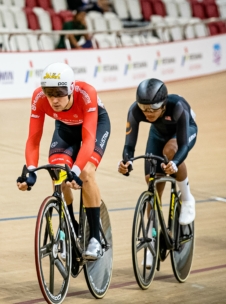 Zwei Radfahrer fahren ein Rennen auf einer Indoor-Bahn. Der vordere Radfahrer trägt ein rot-weißes Trikot mit Österreich am Bein und einen weißen Helm. Der Radfahrer dahinter trägt einen dunklen Anzug und einen schwarzen Helm. Im Hintergrund sind Zuschauer zu sehen.