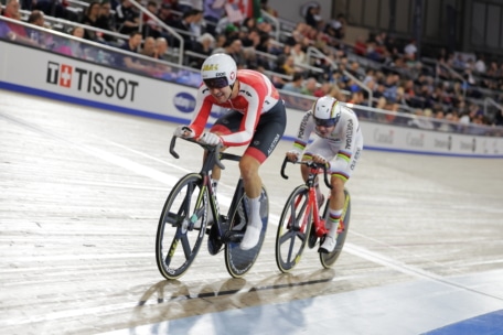 Zwei Radfahrer treten auf einer Indoor-Velodrombahn gegeneinander an, wobei der eine Radfahrer dem anderen dicht auf den Fersen ist. Die Zuschauer schauen von den Tribünen im Hintergrund zu. Beide Fahrer tragen aerodynamische Helme und Rennanzüge.