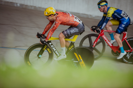 Zwei Radfahrer in Rennanzügen und Helmen fahren auf einer Radrennbahn um die Wette, wobei die Bewegungsunschärfe die Geschwindigkeit betont. Der vordere Radfahrer trägt orange und gelb, der hintere blau und gelb. Grüne Unschärfe im Vordergrund.