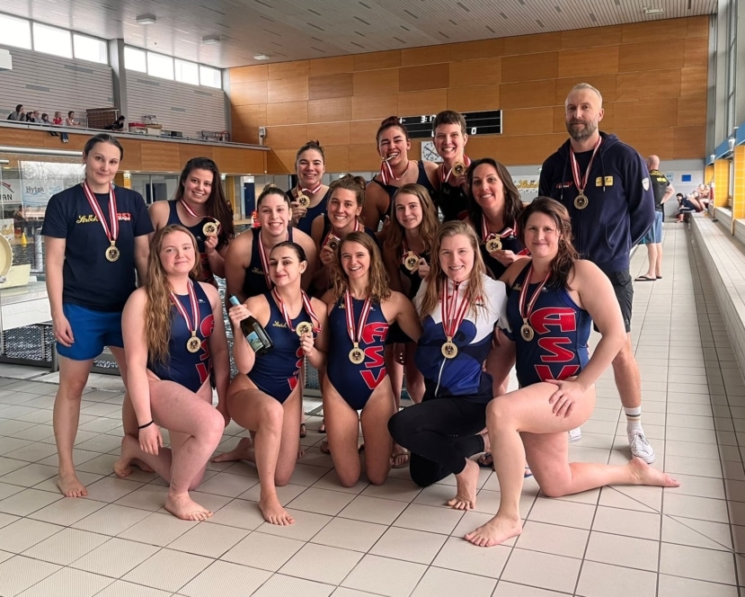 Ein Frauenschwimmteam posiert lächelnd und mit Medaillen in der Hand an einem Schwimmbecken. Die meisten tragen Badeanzüge mit dem Aufdruck ASV. Einige tragen Medaillen um den Hals. Ein männlicher Trainer steht auf der rechten Seite.