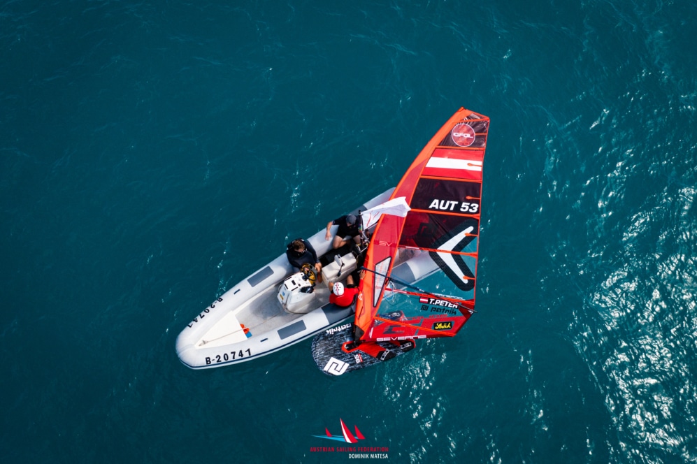 Luftaufnahme eines kleinen Bootes neben einem Windsurfer mit einem roten Segel mit der Aufschrift AUT 53, der auf klarem blauen Wasser treibt. Zwei Personen befinden sich in dem Boot und eine auf dem Windsurfbrett.