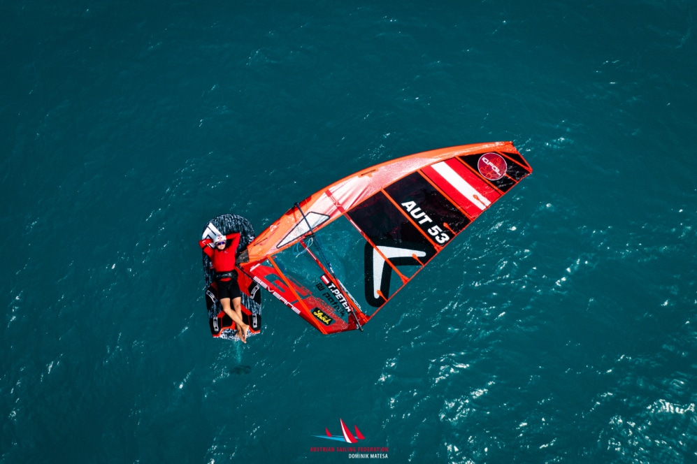 Eine Person in roter Ausrüstung beim Windsurfen auf tiefblauem Wasser, die sich an einem großen roten und orangefarbenen Segel mit der Aufschrift AUT 53 festhält. Das Bild ist von oben aufgenommen und zeigt deutlich das Brett und das Segel.