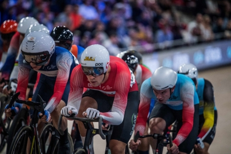 Eine Gruppe von Radfahrern in aerodynamischen Helmen und farbenfrohen Rennanzügen fährt auf einer Indoor-Velodrom-Strecke um die Wette, während die Zuschauer im Hintergrund verschwimmen.