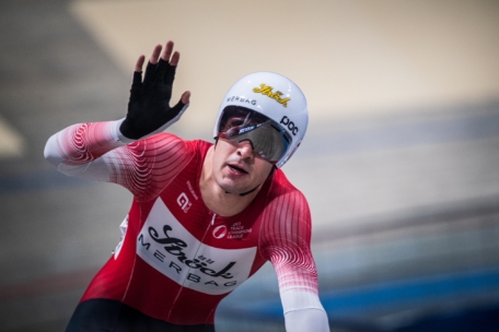 Ein Radfahrer in einem rot-weißen Rennanzug und Helm hebt eine Hand, während er auf einer Indoor-Bahn fährt. Er trägt eine Sonnenbrille und scheint in Bewegung zu sein, möglicherweise während eines Wettkampfs oder einer Veranstaltung.