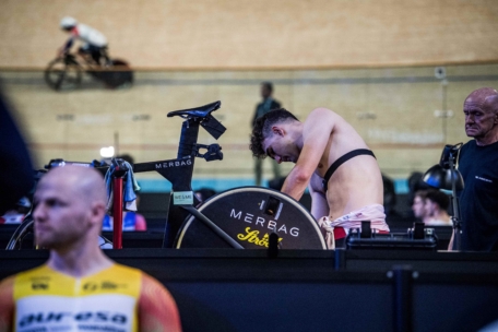 Ein Radfahrer in Radlerhosen und mit Brustgurt lehnt sich über sein Fahrrad und sieht müde aus, während er sich in einem Indoor-Velodrom befindet. In der Nähe sind andere Radfahrer und Fahrräder zu sehen, und im Hintergrund fährt ein Radfahrer auf der Bahn.