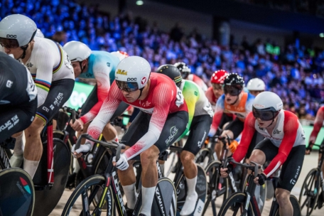 In einem überdachten Velodrom, umgeben von zahlreichen Zuschauern und unter heller Beleuchtung, liefern sich Radfahrer in Helmen und Rennanzügen einen engen Wettkampf.