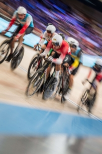 Eine Gruppe von Radfahrern, die Helme und Rennanzüge tragen, rasen um eine Indoor-Bahn, aufgenommen mit Bewegungsunschärfe, um ihre schnelle Bewegung und Intensität zu betonen.