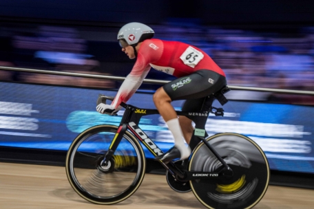 Ein Radfahrer in einem rot-weißen Trikot fährt in einer Halle mit hoher Geschwindigkeit auf einem Rennrad, wobei ein unscharfer Hintergrund die Bewegung unterstreicht.