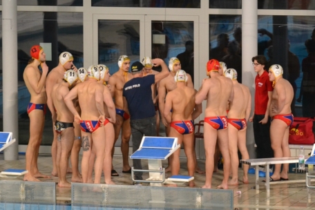 Eine Herren-Wasserballmannschaft mit Badekappen und Badeanzügen versammelt sich am Schwimmbecken und hört ihrem Trainer zu. Zwei Mitarbeiter in roten Outfits stehen in der Nähe. Die Szene spielt sich in einem Innenraum vor großen Fenstern ab.