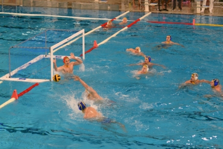 Ein Wasserballspiel ist im Gange. Ein Spieler wirft den Ball in Richtung Tor, während der Torwart mit roter Mütze eine Hand zum Abwehren hebt. Andere Spieler mit blauen und gelben Mützen schwimmen im Becken und schauen zu.