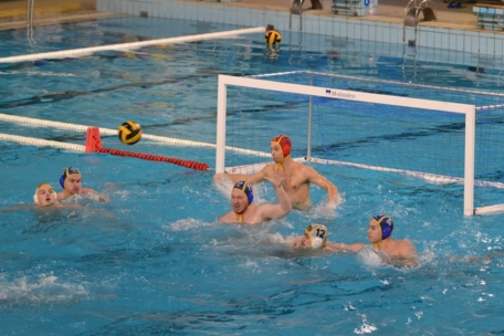 Wasserballspieler treten in einem Schwimmbecken in der Nähe eines Tores gegeneinander an. Der Torwart mit der roten Mütze ist so positioniert, dass er den Ball abblockt, während die Spieler mit blauen und gelben Mützen schwimmen und nach dem Ball greifen. Die Szene ist actionreich und wettbewerbsorientiert.