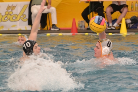Zwei Wasserballspieler treten in einem Schwimmbecken gegeneinander an; der eine trägt eine weiße Kappe mit der Nummer 8 und hält den Ball, während der andere mit einer dunklen Kappe mit der Nummer 10 mit erhobenem Arm verteidigt. Im Hintergrund sitzen Zuschauer.