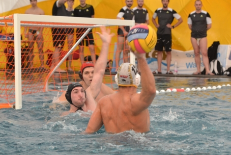 Ein Wasserballspieler bereitet sich darauf vor, den Ball in Richtung Tor zu werfen, während ein Verteidiger den Ball abblockt und der Torwart sich darauf vorbereitet, den Schuss zu stoppen. Im Hintergrund stehen mehrere Personen auf dem Pooldeck.